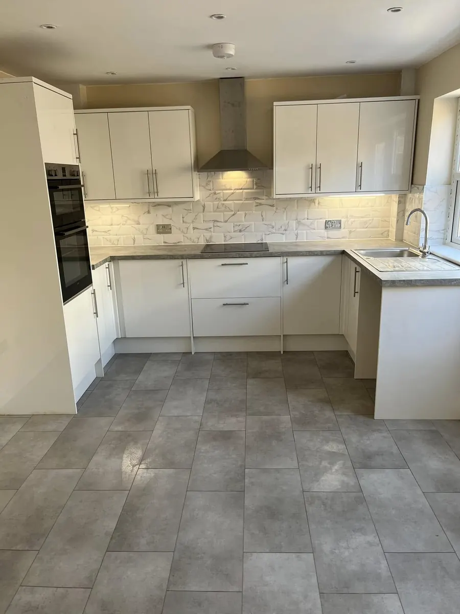 White gloss kitchen installation with marble-effect metro splashback tiles, stainless extractor hood, built-in double oven and grey tiled floor